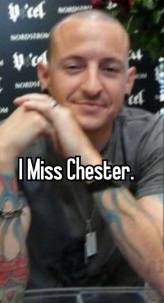 a man sitting at a table with tattoos on his arms and chest, in front of a sign that says i miss chester