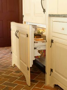 an open cabinet in the middle of a kitchen with red brick flooring and white cabinets