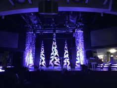 the stage is lit up with purple lights and tall pillars in front of it, while people are seated at tables behind them