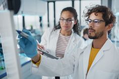 two people in lab coats and gloves looking at something on a white board while another person holds a clipboard