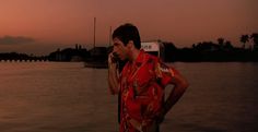 a man standing next to a body of water while talking on a cell phone with the sun setting in the background