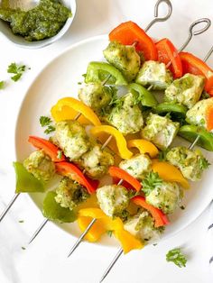 a white plate topped with chicken and veggies on skewers next to a bowl of pesto