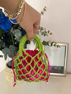 a hand holding a crocheted purse next to a vase with flowers in it