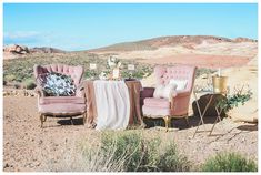 two pink chairs and a table with flowers on it in the middle of an arid area