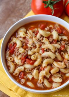 a white bowl filled with pasta and meat soup next to tomatoes on a yellow napkin