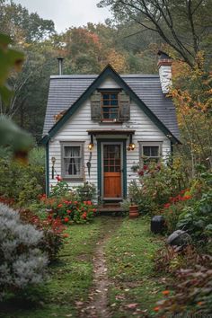 a small white house in the woods with flowers and trees around it's front door