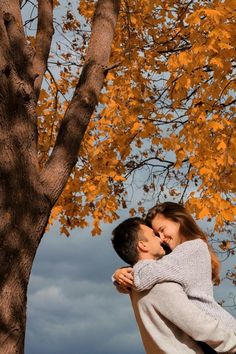 a man holding a woman in his arms under a tree with yellow leaves on it