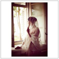 a woman in a wedding dress standing by a window with her veil pulled back and looking out the window