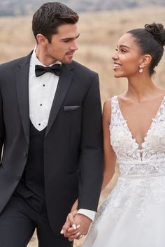 a bride and groom hold hands as they walk through an open field in their tuxedos