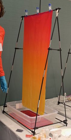 a woman in an orange shirt and blue gloves stands next to a painting easel