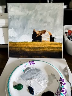 a white plate with some paint on it next to a painting and brush in the foreground