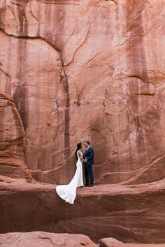 a bride and groom standing in front of a rock formation at the base of a cliff