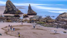 several people are standing on the beach near some rocks and water, while one person is drawing something in the sand