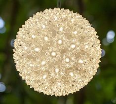 a white ball hanging from a string in front of some green leaves and trees with lights on it