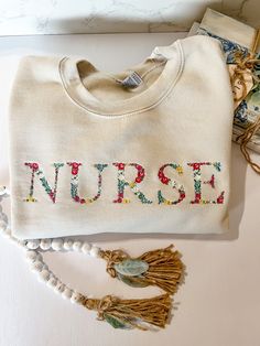 This embroidered floral font is absolutely beautiful. The detail to the flowers and colors are stunning.  Nurse embroidered to the front. Approximately 11x2 inches. Embroidered Long Sleeve Sweatshirt For Spring, Spring Sweatshirt With Letter Embroidery And Long Sleeves, Spring Long Sleeve Sweatshirt With Letter Embroidery, Long Sleeve Sweatshirt With Letter Embroidery For Spring, Cream Sweatshirt With Embroidered Text For Fall, Spring Cotton Sweatshirt With Floral Embroidery, Cream Cotton Sweatshirt With Letter Embroidery, Spring Floral Embroidered Crew Neck Sweatshirt, Floral Embroidery Cotton Sweatshirt For Spring