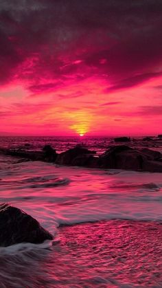 the sun is setting over the ocean with rocks in the foreground and waves on the shore