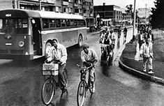 black and white photograph of people riding bikes down the street with buses in the background