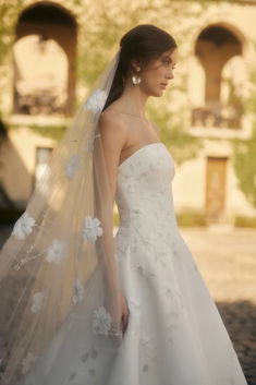 a woman wearing a wedding dress and veil with flowers on the skirt is standing in front of an old building