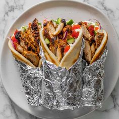 three hot dogs wrapped in tin foil on a white plate next to a marble counter top