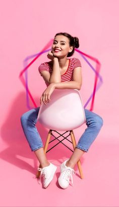 a woman sitting on top of a white chair in front of a pink wall and holding her hand under her chin