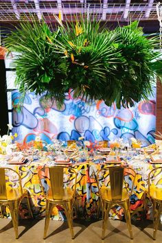 a table set up for a party with yellow chairs and tropical plants hanging from the ceiling