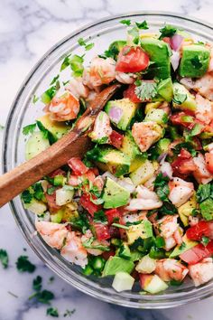 a glass bowl filled with shrimp, avocado and tomato salad next to a wooden spoon