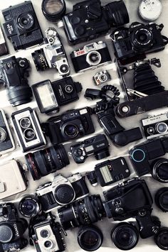 many different types of cameras sitting on top of a white tablecloth covered in papers