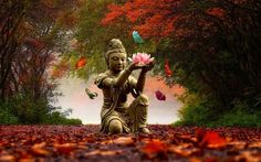 a buddha statue sitting in the middle of a leaf covered forest holding a pink flower