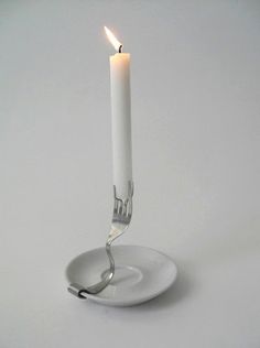 a single white candle on a plate with a fork sticking out of the center, in front of a white background