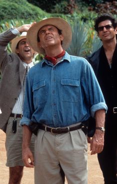 three men standing next to each other in front of trees and bushes, one wearing a cowboy hat