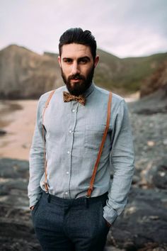 a man with a bow tie and suspenders standing on the beach looking at the camera