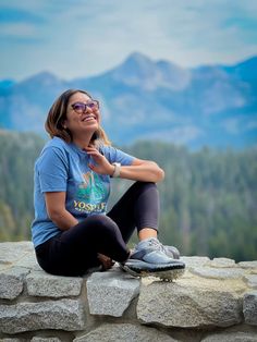 a woman sitting on top of a stone wall with mountains in the background and trees