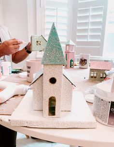 there is a small white birdhouse on the table with people around it and paint supplies