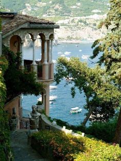 a house on the side of a hill with trees and boats in the water behind it