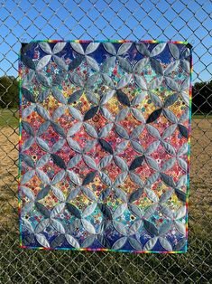 a quilt hanging on a chain link fence in front of a grassy field and blue sky