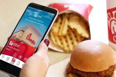 a person holding a cell phone next to a hamburger and french fries