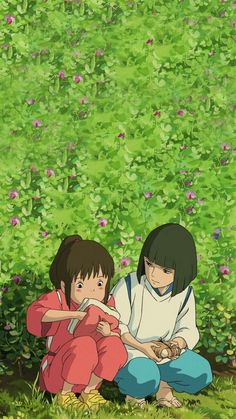 two people sitting on the ground in front of some bushes and flowers, with one person holding