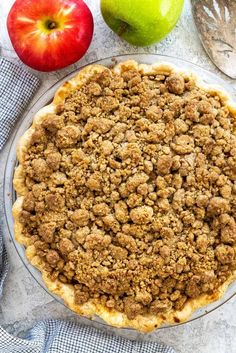 an apple pie with crumbs on top and two apples in the back ground