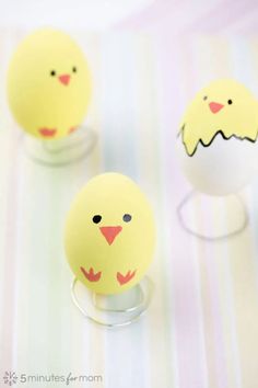 three yellow eggs with faces on them sitting in front of a striped table cloth and one has an egg shaped like a chicken