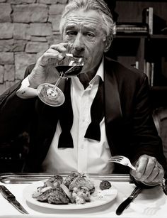 a man in a tuxedo drinking wine at a table with food and utensils