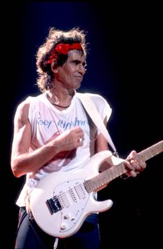 a man in white shirt playing guitar on stage with red bandanna around his head