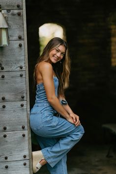 a woman in overalls leaning against a wooden wall with her hand on the leg