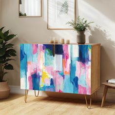 an abstract painting on a cabinet in a living room with potted plants and framed pictures