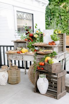 an outdoor table with fruits and vegetables on it, in front of a white house