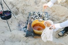 two people are cooking food over an open fire on the beach with their feet in the sand