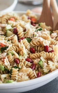 a pasta salad in a white bowl with wooden serving utensils on the side