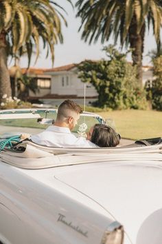 a man and woman sitting in the back of a white convertible car with palm trees behind them