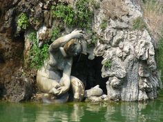 there is a statue in the water next to a rock formation with plants growing out of it