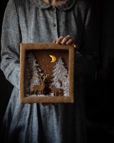 a person holding up a small wooden frame with deer and pine trees on it in the dark