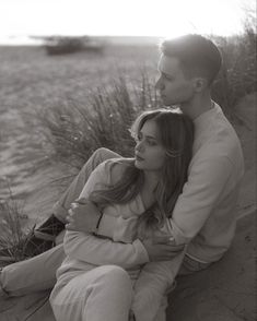 a man and woman sitting next to each other on the sand near some tall grass